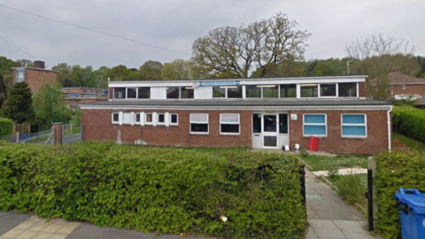 A Google Street View of a building behind a hedge on a cloudy day.