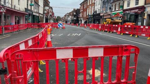 Bright red and white traffic barriers can be seen blocking a high street road from motorists with shops lining each side of the street.