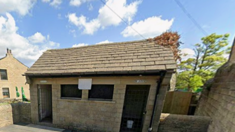 The image shows a small, single‑storey stone building with a pitched roof of grey stone slates and a dark gutter running along the front edge. There are two dark, rectangular window openings. At either end are doorways. Above each doorway are small signs, 'Ladies' and 'Gents'.