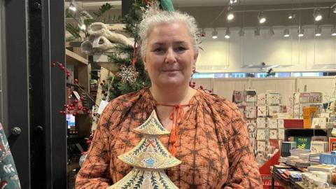 A with with grey hair and an orange top stood in a gift shop and smiling at the camera. She is holding a small wooden Christmas tree