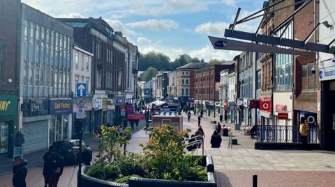 Shops in Walsall town centre and the view includes shoppers on a pedestrianised area and shops on both sides of the street. There is a large plant area in the centre and some individual retail areas. 