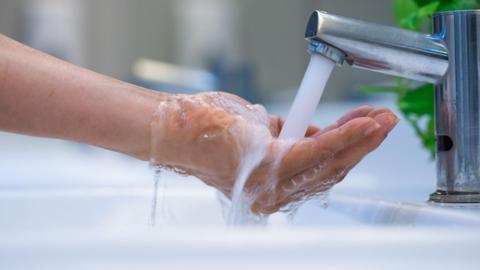 A close up of hands cupped under a silver tap as water streams into them, spilling over into the sink below