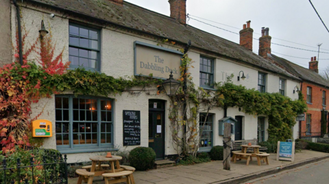 A cream-coloured two-storey public house with wisteria growing across it, level with the pale blue sills on the first floor windows. A sign on the wall says The Dabbling Duck in pale blue. There are two round wooden picnic tables either side of the front door, which is dark green. A blackboard to the left of the door has unreadable white writing on it, headed Opening Hours
