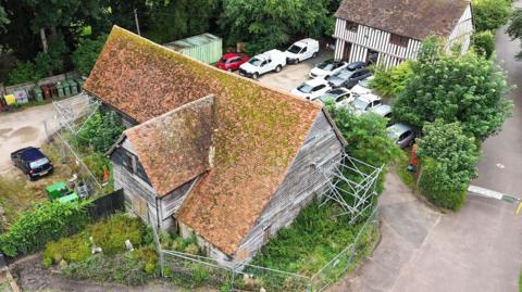 An aerial view of the barn. It is surrounded by fences and scaffolding, and overgrown shrubbery. There are some cars parked between the barn and another half timbered Tudor house. The roof is red with lots of moss growing on it. 