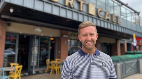 Adam Dickman stands in front of a pub he owns in Sunderland city centre called Port of Call. He is wearing a navy blue and white striped polo shirt. 