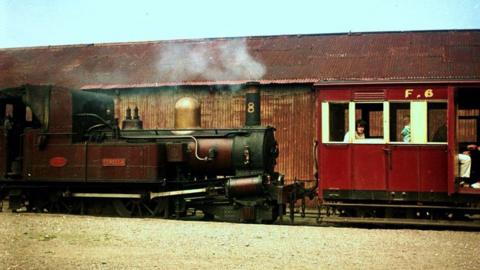 An old photo of the F6 coach being used, it is red with white window frames. A handful of passengers can be seen in the carriage, which is attached to a steam train, with smoke coming from it. 