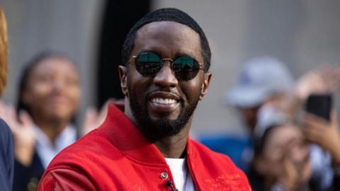 Sean Diddy Combs sits for an interview. He is wearing a red jacket, white T shirt and black sunglasses. He is smiling. People can be seen in the background taking pictures and smiling.
