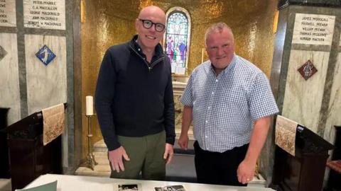 A bald man with glasses and a navy quarter-zip standing next to a shorter man with short white hair wearing a blue and white chequered shirt.
