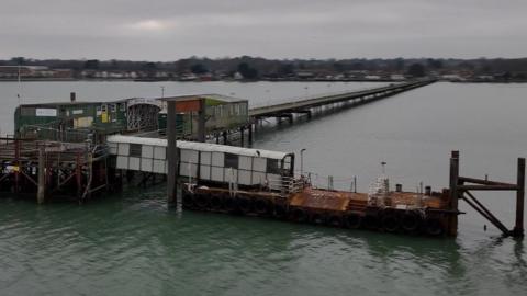 Image is taken by a drone and shows the length of Hythe pier, surrounded by water. At the end is the waiting area and the floating pontoon which is used by the Hythe ferry as a landing stage. 