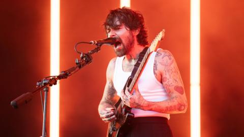 Simon Neil of Biffy Clyro onstage -he is wearing a vest while playing the guitar and singing into a microphone.

He is bearded and has tattoos over his arms.