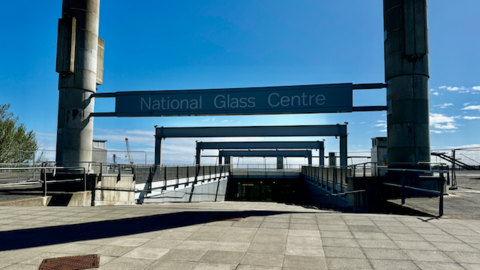 National Glass Centre in Sunderland. Two concrete pillars stand on either side of the centre's wide entrance, which runs into the ground. A sign with the centre's name hangs between the two pillars.