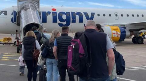 A group of people wait on an airport runway next to a clear tunnel leading up to a plane which says 'Aurigny' in blue writing.