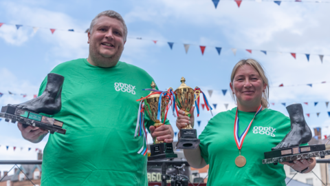 Toe wrestling champions, Ben Woodroffe, also known as "Toe-tal Destruction" and Lisa Shenton, whose wrestling name is "Twinkletoes". The pair wear green T-shirts and hold trophies in their hands.