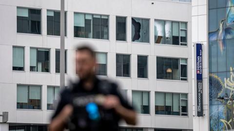 An image showing a building with two broken windows shattered by gunfire. In the forefront is a blurred outline of a Montreal police officer.