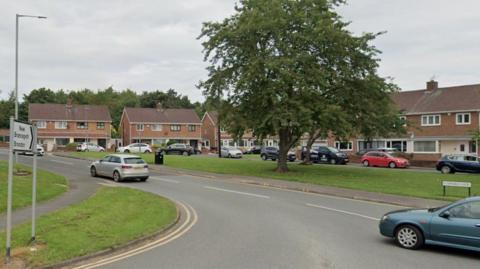 A road with two cars driving past and a sign on a green reading Sawmills Lane. A tree is growing in the middle of the area of grass and residential houses surround the area.
