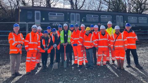 Construction workers, dressed in hi-vis and hard hats, standing at the railway construction site. A green GWR rain is behind them. The group is made up of 18 people.