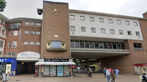 There are people walking past a red brick building with a grey-painted front in a pedestrianised shopping precinct. The word "Hertford Street" can be seen on a sign.

