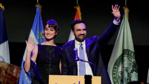 Zohran Mamdani waving at a podium with flags behind him and a dark background, his wife Rama Duwaji next to him 