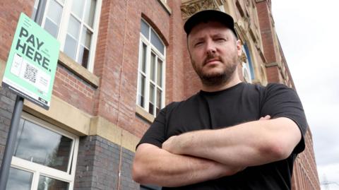 Steven Shepherd, who has brown stubble and is wearing a black baseball cap and black t-shirt, stands next to a sign saying 'Pay Here' with his arms folded and a stern expression. 
