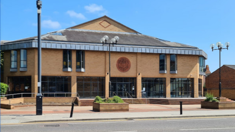 A photo of the modern brick building which has extensive black windows on the ground floor and a pitched roof.  Steps surround the building and a CCTV pole is in the foreground of the picture.