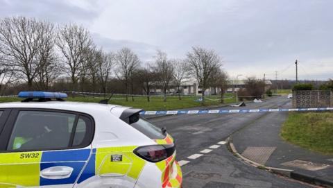 A police car is in the foreground on a road. Police tape is in the background.