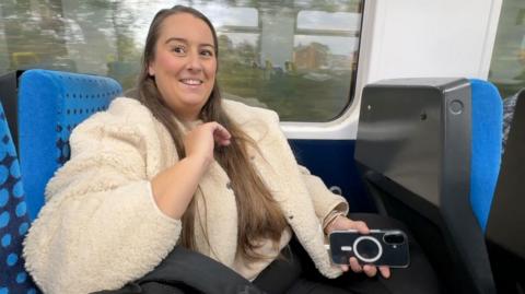 A woman in a warm-looking cream coat sits in a blue Northern Rail train seat. She has long brown hair, and is smiling. She's holding a black mobile phone in her left hand. The train windows behind her reflect the windows on the other side of the carriage.