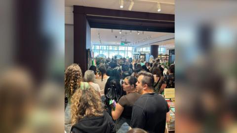 A picture inside a large bookshop at the back of a large crowd, which stretches to the far, windowed wall.
