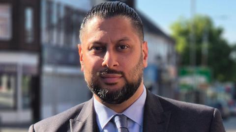 Shaz Saleem facing the camera. He has black hair and a black beard and moustache. He is wearing a grey suit with a blue shirt and tie.