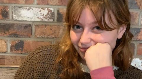 Kat Nellist is looking at the camera while resting her face against her closed fist while her elbow rests on the table. She has shoulder-length wavy auburn hair and curtain bangs. She is wearing a brown knitted jumper and is sitting in front of a brick wall