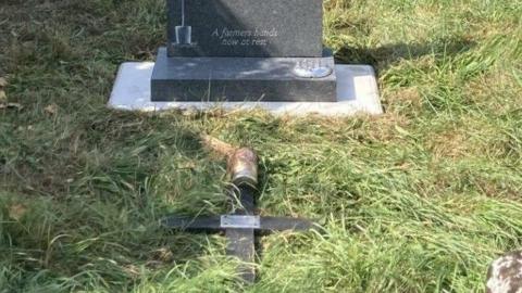The wooden cross is lying face down in grass in front of a grey memorial stone. At the base the words are carved: A farmer's hands now at rest.