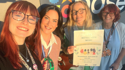 Four women standing in front of a sign that reads 'the Base'. Luiza, second from the right, has blonde hair and glasses. She is holding a poster for the group that bears its name, and the phrase "No pressure, just come as you are"