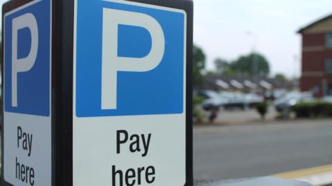 A pay and display machine is pictured in a car park. A large P in a white font is painted on the sign on a blue background. The words, 'Pay here' are also 