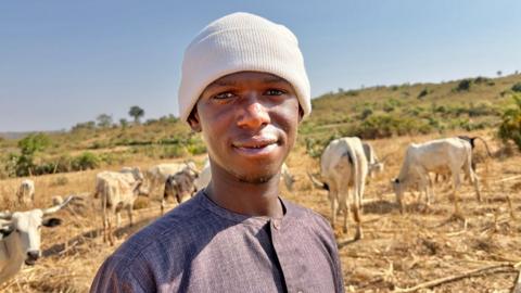 Aliyu Abdullahi Isa, a Fulani herder wearing a white knitted hat. Behind him are white cattle grazing