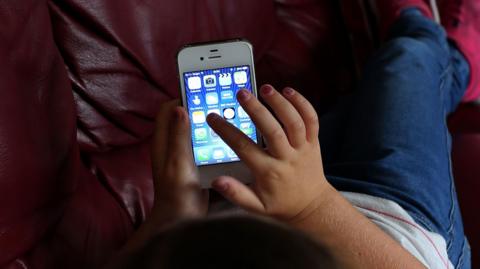 A child with sparkly fingernails, a white top, jeans and pink trainers is tapping a modern mobile phone. The child's face is cut out of shot as they lay on a red leather settee with their feet up.