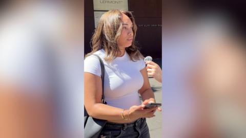 A young woman wearing a white t-shirt looks at someone off-camera and holds her phone