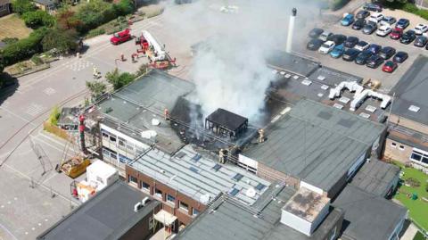 An aerial photograph of Ormiston Cliff Park Primary Academy on Orde Avenue in Gorleston. Fire service members are seeing working on the roof which is charred and has smoke billowing out of it. 