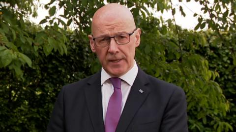 First Minister John Swinney stands in front of shrubbery wearing a dark suit with a purple tie and light shirt