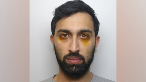 A man with dark hair parted to one side. He also has a short beard and a trimmed moustache. He has yellow marks around his eyes.