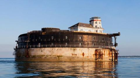 Spitbank Fort is a circular stone fort built in the middle of the sea. It has a railing going round the circumference, and a tall white building on one side. The photo is taken at either sunrise or sunset, with the fort bathed in gold light.