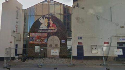 A Google Street View screengrab of the façade of the former club, building painted white with a glass column and 'Club Heaven & Hell' sign with an illustrated angel and devil. The building is in poor condition and has metal security fencing surrounding it with 'Manor' branded signs on.