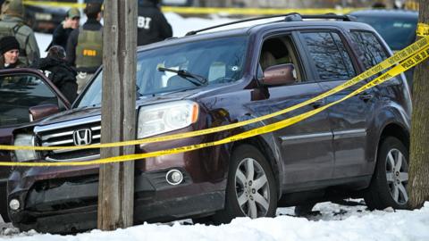 The car in which the woman was driving is seen crashed into a lampost with police tape in the foreground and a bullet hole visible in the windshield