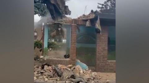 An excavator with a large pneumatic drill attachment breaks up a brick bus shelter. It is working on the roof and a mural on a back wall can just be seen through the dust.