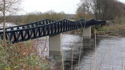 Artist impression of the new bridge, spanning the river and consisting of two concrete posts supported by black steel work with V-shaped trusses and a scalloped rail