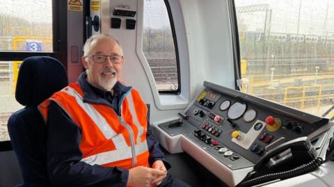 Don Coffey sat in the train driver cabin with a dashboard in front of him. He is wearing an orange high visibility jacket.