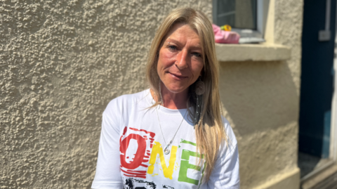 A blonde woman standing in front of a light yellow wall. She's wearing a white T-shirt with the word 'one' written on it.  