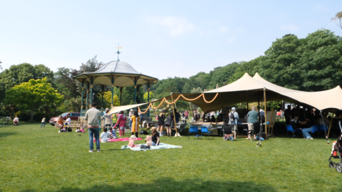 A view of a small canvas stretch tent on grass with a workshop happening underneath it in front of a band stand in Grove Park. There are a number of people wandering about and some on blankets on the grass.