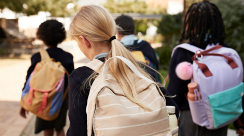 School pupils carrying backpacks are walking along a pavement, away from the camera. They were wearing school uniforms.