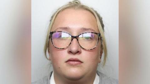 A mugshot of a woman with blond hair in a ponytail, wearing a grey jumper, and tortoiseshell glasses.