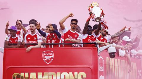 Bukayo Saka of Arsenal (R) lifts the English Premier League trophy as he and teammates ride an open-top bus during Arsenal Champions Parade in north London