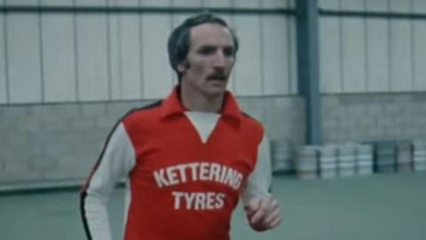 Derek Dougan, who has a moustache and receding hairline, is wearing a red football shirt with white sleeves. "Kettering Tyres" is emblazoned across the front in white capital letters. He appears to be training in a large sports hall and there are what appear to be beer kegs on the wall behind him.